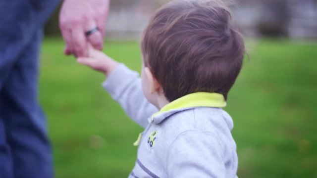 Adorable little boy holding his dad's hand while walking in a park, then looking behind him