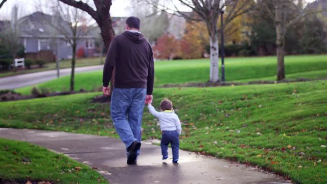 Adorable little boy holding his dad's hand while walking in a park, mom following with a stroller