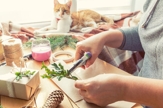Woman wrapping eco Christmas gifts presents with brown paper, string and natural branches and decor elements on wooden table at home
