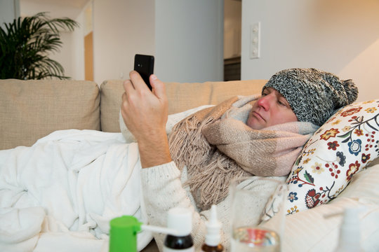 Sick Man Wearing Scarf Lying On Couch At Home Under A Blanket Checking Temperature. Browsing With Smart Phone. Living Room With Table Full Of Medicine And Pills. Man With Running Nose And Cough. 