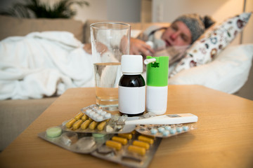 Fototapeta na wymiar Sick man wearing scarf lying on couch at home under a blanket checking temperature. Living room with table full of medicine and pills. Man with running nose and cough. 