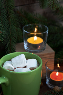 Hot chocolate with marshmallow in green cup and two light candles with Christmas and New Years decor and Christmas tree on dark wooden background