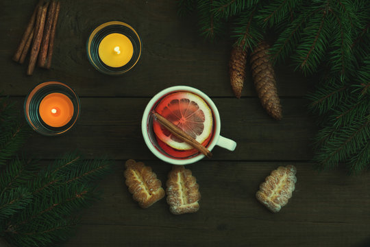 Hot tea with lemon and cinnamon in white cup. Two light candles, Christmas tree and cookies on dark wooden background. Top view.