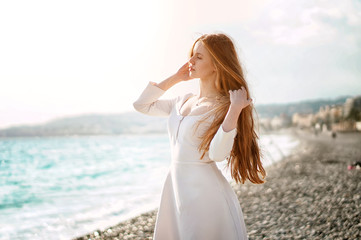 Fototapeta na wymiar Outdoor summer portrait of young pretty woman with great hair looking to the ocean at europe beach, enjoy her freedom and fresh air, wearing stylish white dress.