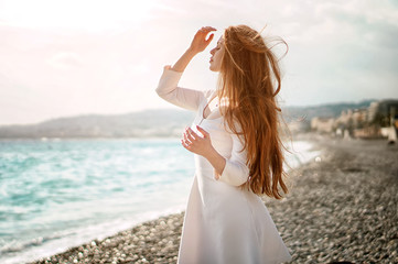 Outdoor summer portrait of young pretty woman with great hair looking to the ocean at europe beach, enjoy her freedom and fresh air, wearing stylish white dress.