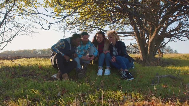 Four multiracial friends sitting on ground near beautiful tree and sharing photo images on smart phone. Round camera, backlit.