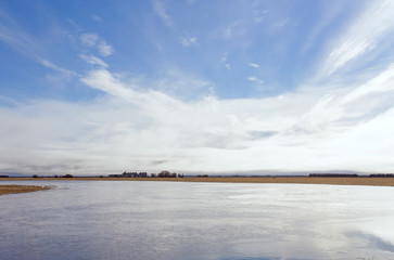 A harvested field flooded with water with a farm yard in the distance under cloudy sky in rural landscape
