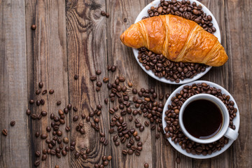Breakfast coffee - cup and croissant with coffee beans on wooden background.