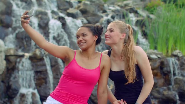 Girls selfie. Girlfriends taking selfie with smartphone outdoors. Selfie women. Multi ethnic girls taking selfie in park. Cheerful friends taking self photo with iphone near waterfall
