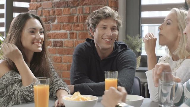 Group of young friends talking around a table with healthy orange drinks in a leisure day in modern loft interior. Happy young college students having spending great time together.