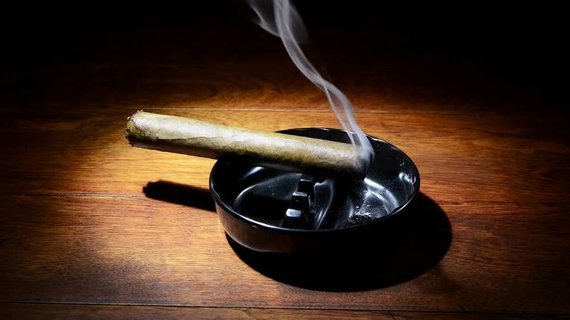 A burning cigar in a classic black ashtray streaming smoking in a dark, moody setting.