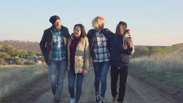Slowmotion of happy diverse group of four young adult men and women in coats walking on dirt path outside during late afternoon
