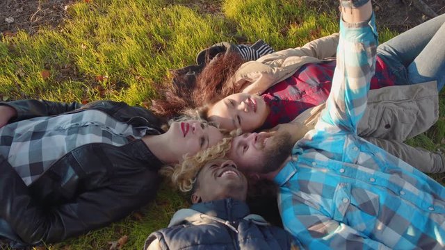 Diverse quartet of friends taking a self portrait with their camera phone while laying down on grass outside. Top view