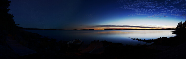 Panorama of a lake at dusk and after nightfall. On the right side sky and lake are illuminated by the sunset. On the left side stars are visible. The foreground is the dark silhouette of the coastline