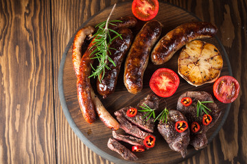 Close-up photo of mixed grilled meat platter. Beef, pork, poultry, sausages, grilled garlic, chili pepper, red tomatoes on wooden rustic background.