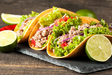 Photo of Mexican tacos with ground meat, beef, beans, onions and salsa on wooden background. Ketchup sauce and lime. A glass o beer in the background.