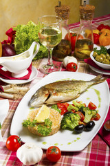 Fried sea bream on a plate with a side dish of broccoli, cherry tomatoes, rice, olives and lemon served beside fresh vegetables, sauce, olive oil and white wine.