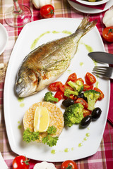 Fried sea bream on a plate with a side dish of broccoli, cherry tomatoes, rice, olives and lemon.