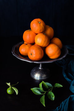 mandaranci accatastati su piatto per dolci in metallo. Piccolo mandarino acerbo e ramo con foglie fuori fuoco in primo piano