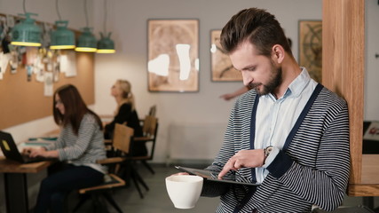 young handsome bearded man uses touchscreen tablet and drinking coffee in the modern startup office.