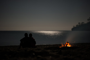 Silhouette of a young couple on summer beach near campfire at mo