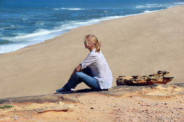 Fototapeta na wymiar Beautiful calm blond woman sitting alone in relax pose on the ocean beach, Nazare, Portugal
