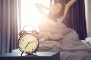 Closeup alarm clock having a good day with background happy woman stretching in bed after waking...