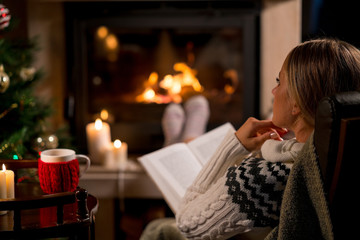 Woman is sitting with cup of hot drink and book near the fireplace