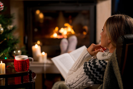 Woman is sitting with cup of hot drink and book near the fireplace