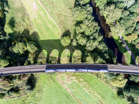 View from above: moving train on the railway bridge with shadow dropping from the bridge over green fields, New Mills, Derbyshire, England
