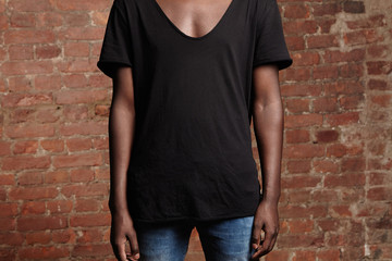 Cropped view of young African man with athletic body dressed in blue jeans and black t-shirt with scooped neck, posing isolated against red-brick wall background with copy space for your information