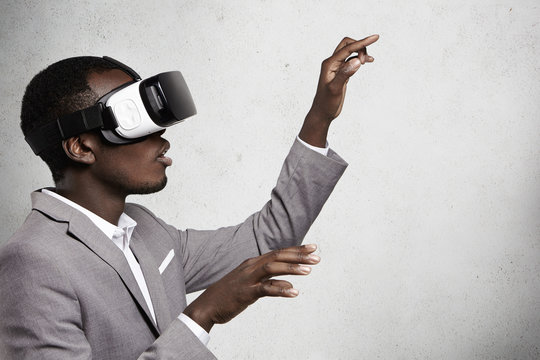 Technology, innovation, cyberspace and gaming. Profile of fascinated dark-skinned businessman in elegant suit wearing oculus rift headset, gesturing as if touching something while playing video games
