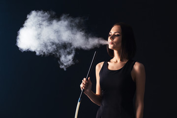 Young, beautiful girl smokes a hookah. It produces smoke from his mouth. Business style clothing. The pleasure of smoking.


