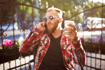 Obraz premium Handsome young man holding mobile phone and drinking coffee while sitting in the rest area of the office. Man dressed in a trendy hipster clothing. 