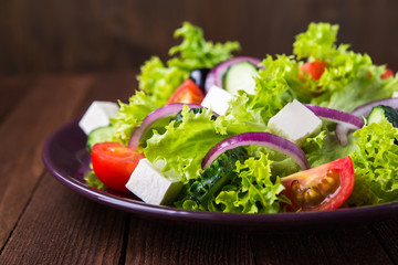 Greek salad (lettuce, tomatoes, feta cheese, cucumbers, black olives, purple onion) on dark wooden background close up. Healthy food.