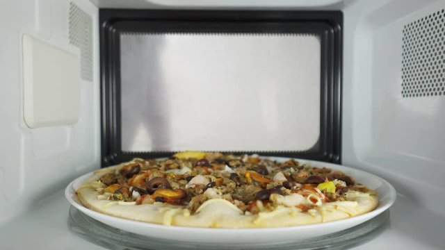 Man reheating baked seafood pizza in the microwave oven. He opens the door of the oven and takes out a dish with hot pizza Frutti di Mare topped with mussels shrimp and olives.