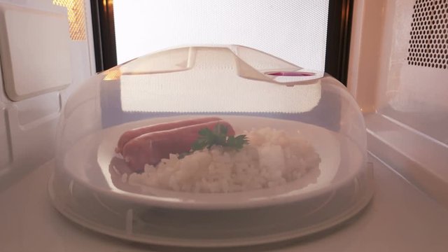  Portion of rice and sausages covered with plastic plate cover heating in the microwave oven inside view