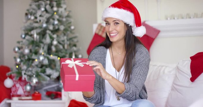 Excited young woman opening a Christmas gift with a beaming smile of anticipation as she sits in her decorated home in Santa hat
