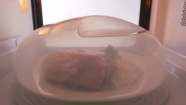 Plate with crispy pan fried chicken leg and rice garnish covered with microwave cover reheating in the microwave oven.