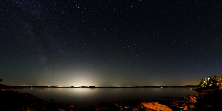 Panorama of the night sky above a lake in Sweden. The Milky Way is visible on the left and the bright light of large cities on the opposite side of the lake. In the foreground is the rocky coastline.