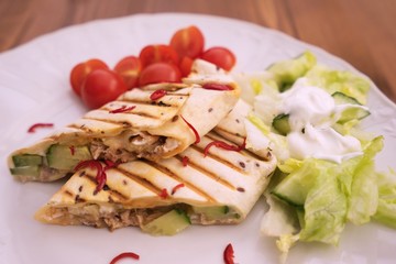 Detailed Picture of baked wraps, fachitas or burrito with fresh chili pieces, cherry tomatoes and fresh salad with cream served on the white plate and wooden table. Typical example of mexican cuisine.