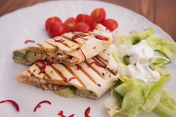Detailed Picture of baked wraps, fachitas or burrito with fresh chili pieces, cherry tomatoes and fresh salad with cream served on the white plate and wooden table. Typical example of mexican cuisine.