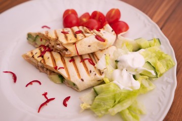 Close up of baked  tortilla wraps, fajitas or burrito with fresh chili pieces, cherry tomatoes and fresh salad with cream served on the white plate and wooden table. Typical example of mexican cuisine