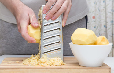Man's hands with a grater grate the potatoes on the wooden board for potato pancakes in the kitchen. Healthy eating and lifestyle.