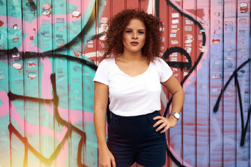 Naklejka premium A red-haired hipster girl wearing white t-shirt and black shorts is looking at camera while standing beside the graffiti wall. An attractive woman with freckle face is standing on the painted wall.