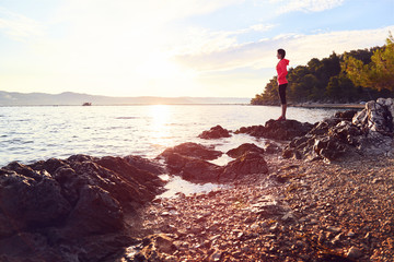 Fototapeta premium Runner resting from routine exercise. Happy jogger relaxing at the beach after run, standing on the stones and looking at the sea. Active young woman enjoying sunrise.