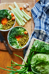 Top view of vegetable mix in bowls with green peas, cucumbers, carrots, lettuce and dill, standing on a wooden table