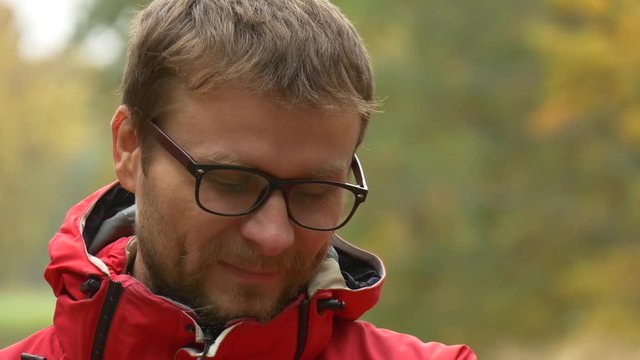 Man in Eyeglasses is Smiling Looking Down Backpacker Outdoors Autumn Park Yellow Trees Behind the Tourist in Red Sporty Jacket Man Resting at the Nature