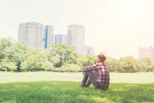 Young Hipster Man Sitting On Grass In The Park Alone Against The