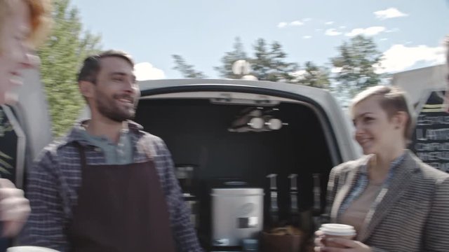 Young ginger man and two friendly women standing before mobile coffee van chatting with barista and drinking coffee